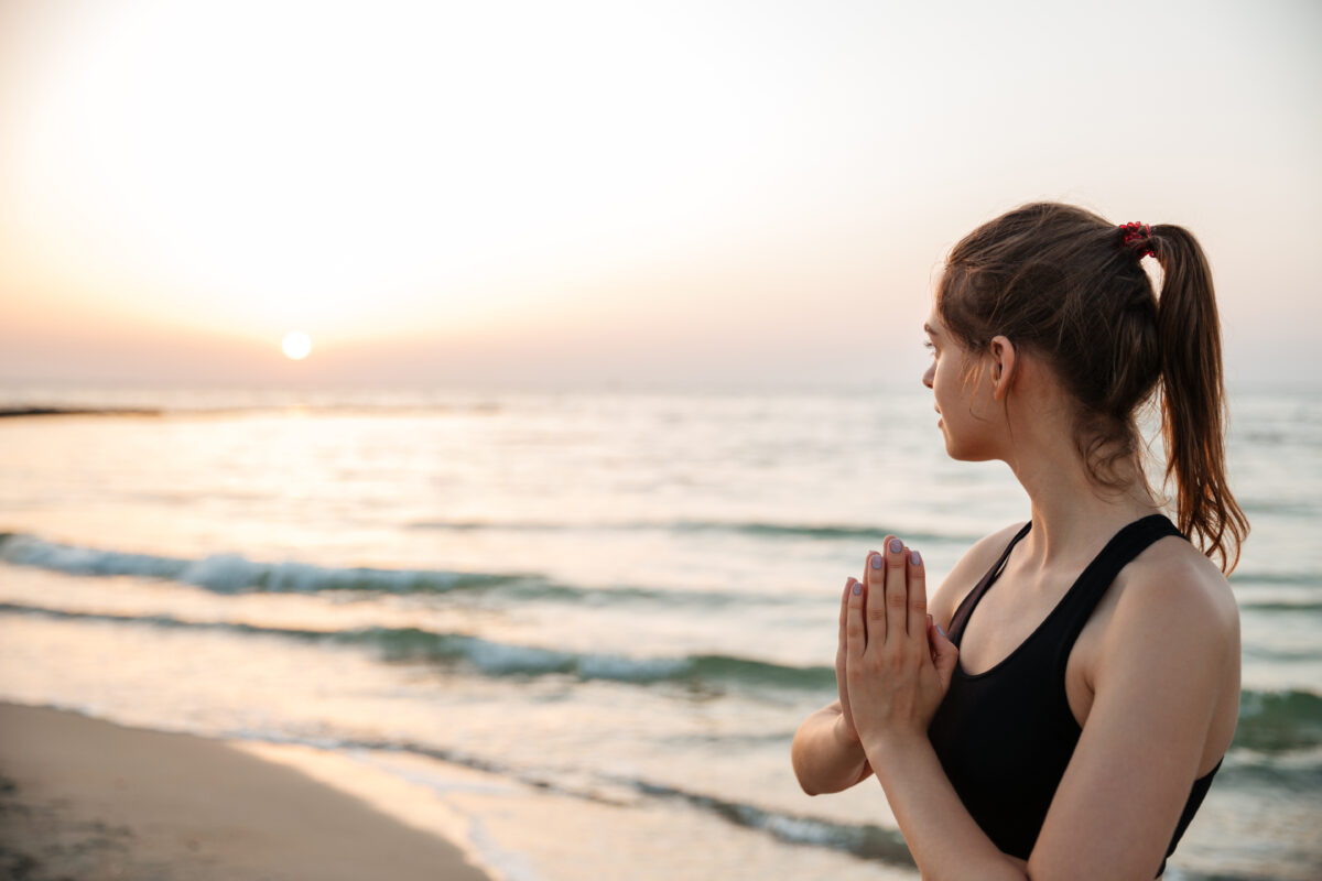 Frau in Yogapositon- Handflaechen aneinander blickt aufs Meer und den Sonnenuntergang am Horizont